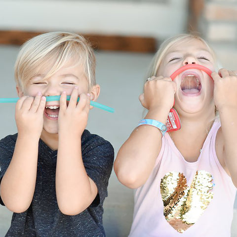 Two kids holding their reusable silicone straws on their upper lip pretending it's a mustache. Both kids are laughing. 