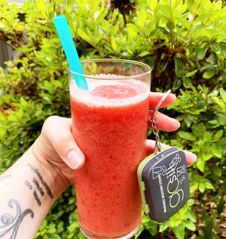 A hand holding a glass filled with a pink smoothie. The glass has a blue reusable silicone straw in it and, with one finger, the hand is holding a keychain travel case for the straw. 