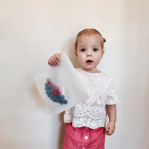 A little girl standing up holding a reusable silicone snack back with her left hand. 