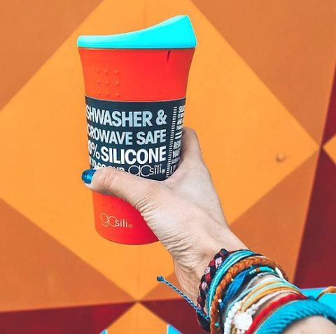 A hand is holding an orange silicone cup with a blue top. The colors match the bracelets and accessories the person is wearing on their wrist. 