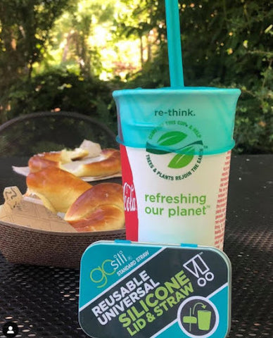 A paper cup with a silicone lid and straw sitting at a table in a patio or park 