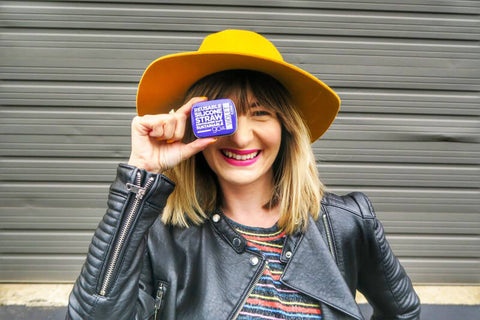 A woman wearing a yellow hat and smiling brightly. She's holding a travel tin for a reusable silicone straw up to her eye. 